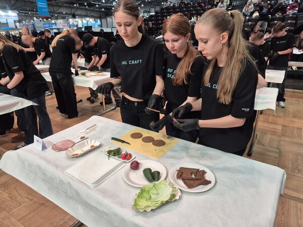 Drei Schülerinnen an einem Tisch mit Zutaten für Sandwiches, darunter Brot, Salat, Gurken und Wurst; im Hintergrund eine Halle mit weiteren Schülerteams und Zuschauern.