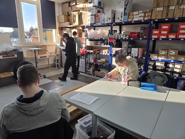 Mehrere Jugendliche arbeiten in einem Lager-/Übungsraum: Zwei Personen prüfen Waren an Regalen, während andere an Tischen sitzen und Unterlagen ausfüllen.