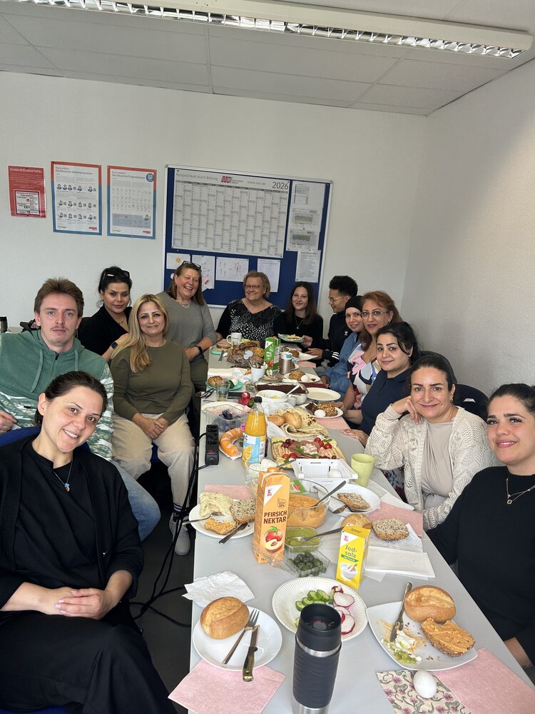 Eine Gruppe von Erwachsenen sitzt gemeinsam an einem langen Tisch bei einem reich gedeckten Frühstück. Auf dem Tisch stehen Brötchen, Aufschnitt, Obst, Gemüse, Getränke und weitere Speisen; die Teilnehmenden blicken lächelnd in die Kamera.