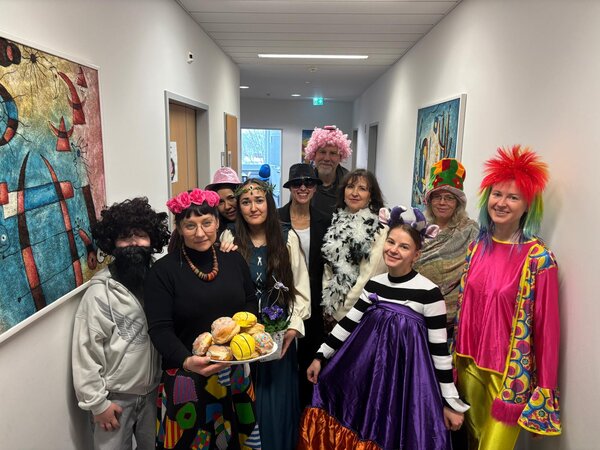 Gruppe von verkleideten Personen in einem hellen Flur, einige mit Perücken, Hüten und bunten Kostümen; vorne hält eine Person ein Tablett mit Berlinern/Krapfen in die Kamera.
