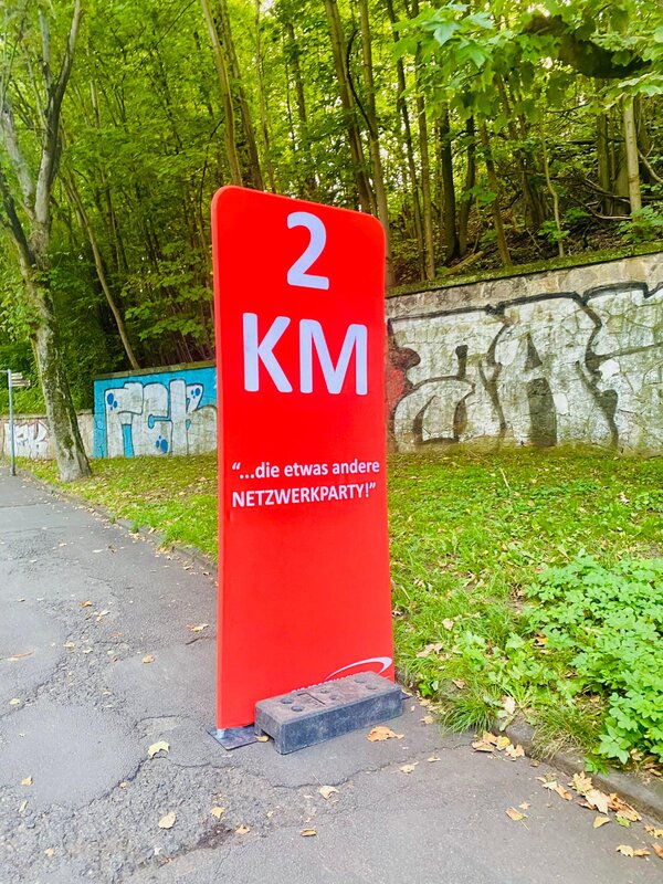 An einer Straße neben einem Waldstück steht ein roter Aufsteller mit der Aufschrift: 2 KM "die etwas andere Netzwerkparty!"