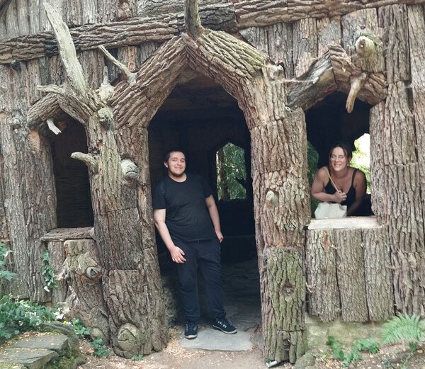 Zwei Personen vor einem fantasievollen, baumstammförmigen Haus mit geschnitzten Details und Fenstern. Eine Person steht im Eingang, die andere schaut aus einem Fenster heraus.