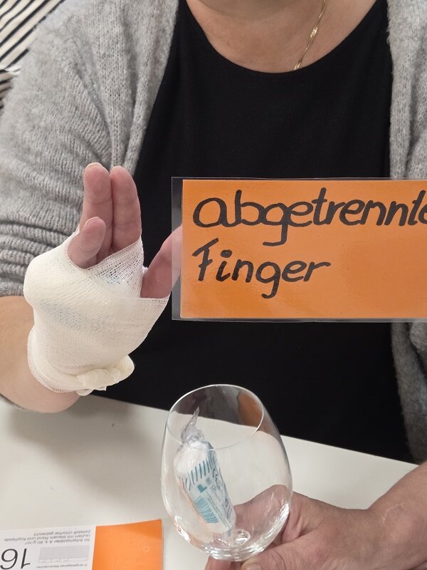Eine Mitarbeiterin mit bandagierter Hand hält ein Schild mit der Aufschrift „abgetrennter Finger“ und demonstriert die richtige Versorgung einer simulierten Verletzung mithilfe eines angelegten Verbands im Erste-Hilfe-Training.