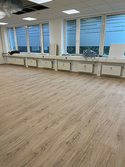 Großer, heller Raum mit frisch verlegtem Holzfußboden, großen Fenstern auf einer Seite und Heizkörpern darunter. Auf der Fensterbank liegen Kabel und Werkzeuge – auch dieser Raum ist im Umbau.