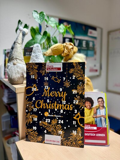 Adventskalender mit der Aufschrift „Euro Schulen – Merry Christmas“ steht auf einem Büroschrank, dahinter dekorative Weihnachtszwerge und eine grüne Zimmerpflanze; rechts daneben ein Prospektständer mit einem Flyer „Euro Schulen – Deutsch lernen“ mit zwei lächelnden jungen Menschen auf dem Titel.