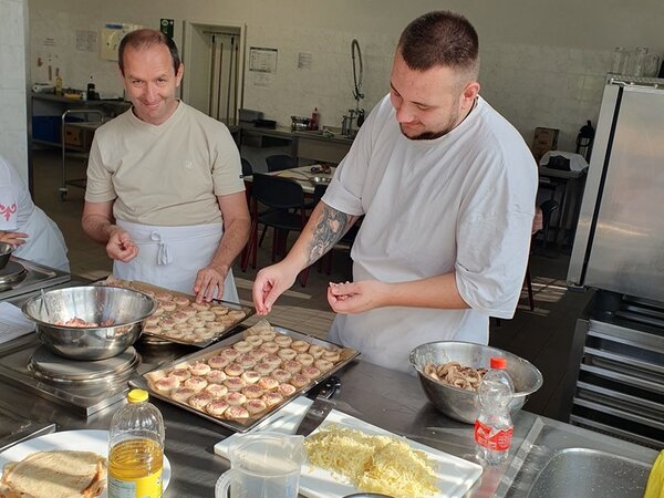 Zwei Teilnehmer stehen in Kochschürze gut gelaunt in der Küche, haben zwei Backbleche köstlicher Speisen vor sich, die sie weiter zubereiten