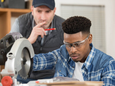Zwei Personen befinden sich in einer Werkstatt. Eine Person bedient eine Kreissäge, während die andere Person zuschaut und einen roten Stift hält. Die Gesichter beider Personen sind aus Datenschutzgründen unscharf.