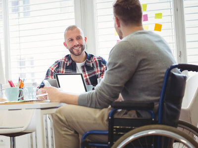 Zwei Männer sitzen an einem Tisch in einem hellen Raum mit großen Fenstern. Der Mann links, der ein kariertes Hemd trägt, lächelt freundlich und hält ein Tablet in der Hand. Ihm gegenüber sitzt ein Mann im Rollstuhl, der ein graues Langarmshirt trägt. Beide wirken entspannt und sind in ein Gespräch vertieft. Auf dem Tisch stehen ein Laptop, ein Notizblock, Stifte und zwei Tassen. Im Hintergrund sind bunte Notizzettel an der Fensterfront zu sehen.
