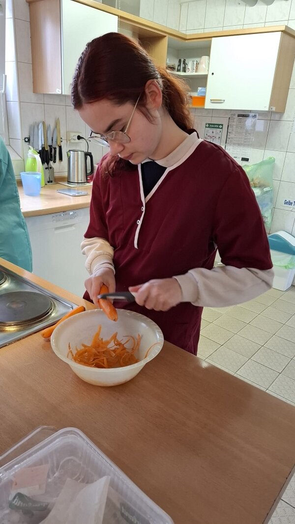 Eine junge Frau mit Brille und roten Ärmeln schneidet Karotten in eine weiße Schüssel in einer Küche.
