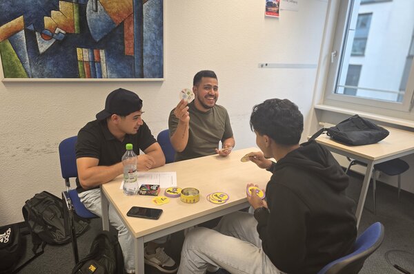 Drei junge Männer spielen ein Kartenspiel an einem Tisch. Einer hält lachend eine Karte hoch, während die anderen aufmerksam zuschauen. Auf dem Tisch liegen Karten und eine Getränkeflasche.