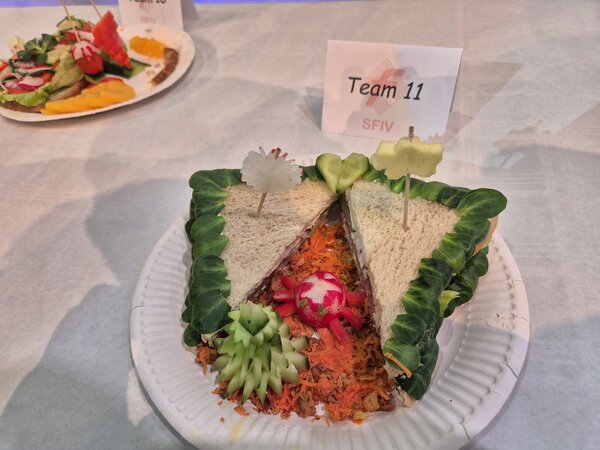Kunstvoll belegte Sandwiches mit Feldsalat und einer Garnitur aus Gurke, Karotte und Radieschen auf einem Pappteller; dahinter ein Schildchen mit der Aufschrift "Team 11".