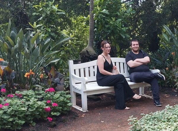 Zwei Personen sitzen auf einer weißen Holzbank in einem üppigen Garten, umgeben von blühenden Pflanzen, Palmen und grünem Laub.