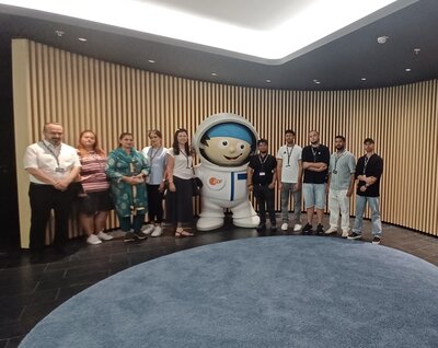 Vor einer Wand aus Holzlamellen steht eine Gruppe von Menschen für ein Gruppenbild mit einer lebensgroßen Mainzelmännchenfigur in einem Astronautenanzug mit ZDF-Logo auf der Brust.
