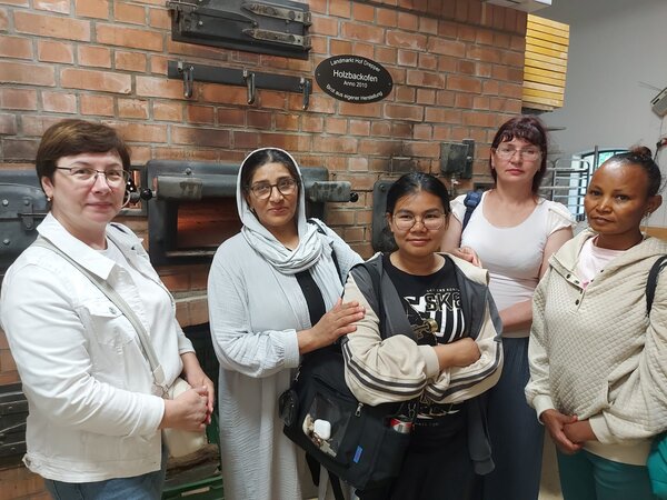 Fünf Frauen stehen vor einer Backofenanlage mit Ziegelwand und blicken in die Kamera. Die Gruppe ist Teil einer Besichtigung oder eines Ausflugs.