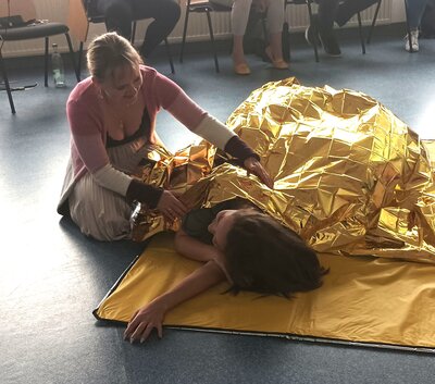 Eine Frau kniet auf dem Boden und hilft einer anderen Person, die unter einer goldfarbenen Rettungsdecke liegt. Die Szene findet in einem Raum mit mehreren Stühlen und Personen im Hintergrund statt. Die Frau unter der Decke liegt auf einer gelben Unterlage.