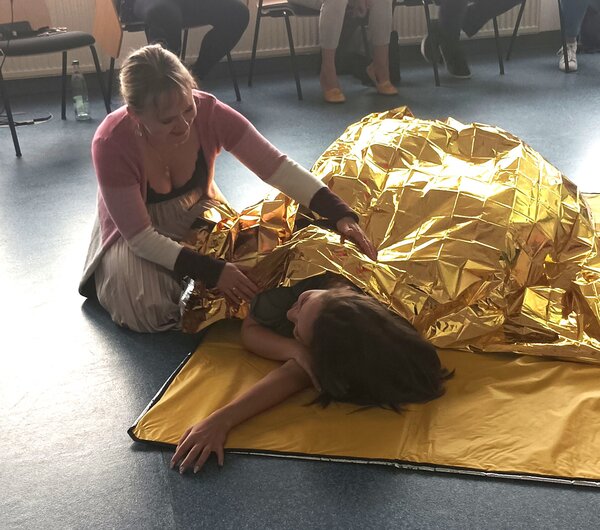 Eine Frau kniet auf dem Boden und hilft einer anderen Person, die unter einer goldfarbenen Rettungsdecke liegt. Die Szene findet in einem Raum mit mehreren Stühlen und Personen im Hintergrund statt. Die Frau unter der Decke liegt auf einer gelben Unterlage.