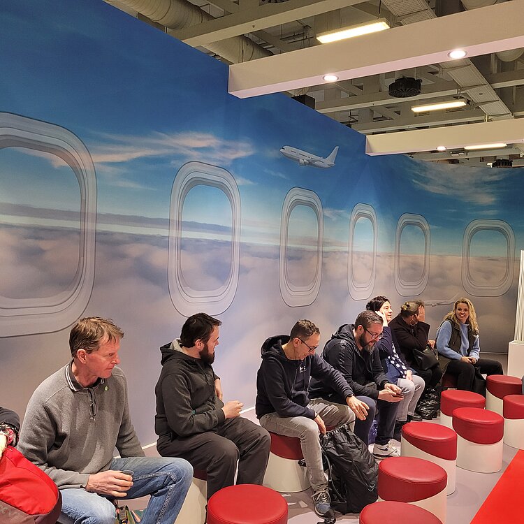 „BO auf Tour – Unternehmen laden ein“ auf der ITB 2025 Menschen sitzen auf runden, roten Hockern in einer Messe-Lounge, die einem Flugzeuginterieur mit Fenstern über den Wolken nachempfunden ist. Ein großes Wandbild zeigt einen Himmel mit einem fliegenden Flugzeug. Die Personen unterhalten sich oder schauen auf ihre Handys.