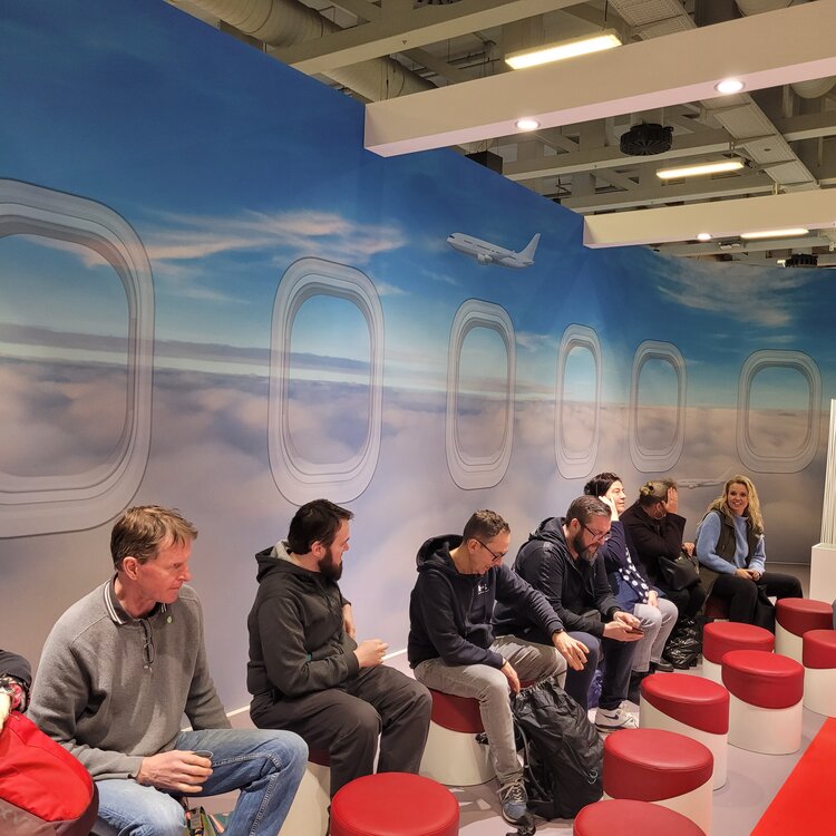 Menschen sitzen auf runden, roten Hockern in einer Messe-Lounge, die einem Flugzeuginterieur mit Fenstern über den Wolken nachempfunden ist. Ein großes Wandbild zeigt einen Himmel mit einem fliegenden Flugzeug. Die Personen unterhalten sich oder schauen auf ihre Handys.