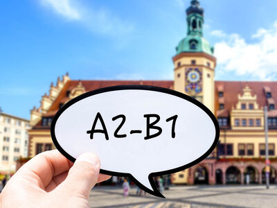Eine Hand hält eine Sprechblase mit der Aufschrift 'A2-B1' in den Vordergrund. Im Hintergrund ist ein historisches Gebäude mit einem markanten Turm und einer Uhr zu sehen. Das Gebäude hat eine charakteristische Architektur mit roten Dächern und einem Platz mit Menschen, die sich in der Umgebung aufhalten.
