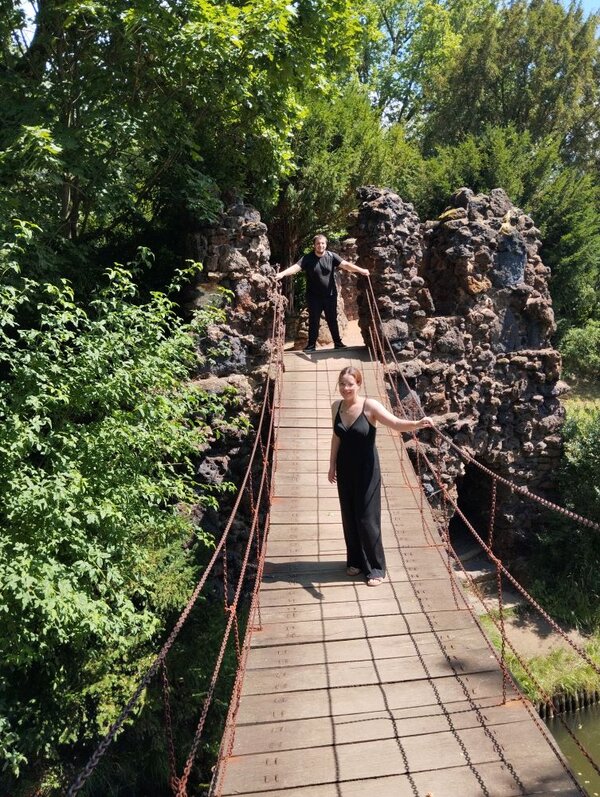 Zwei Personen stehen auf einer hängenden Holzbrücke mit Seilgeländern, die über einen kleinen Bach oder eine Schlucht führt. Eine Person steht vorne, eine hinten. Im Hintergrund sind Felsformationen und dichter Wald zu sehen.