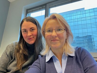 Zwei Mitarbeiterinnen der Euro-Schulen Hannover sitzen im hellen Schulungsraum vor einem großen Fenster und lächeln in die Kamera. Im Hintergrund ist ein modernes Bürogebäude zu sehen. Das Bild entstand während einer polizeilichen Fortbildung zum Erkennen gefälschter Dokumente.