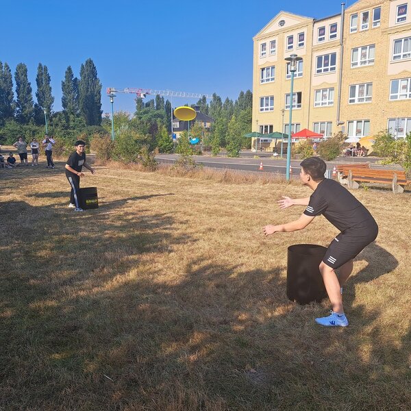 Zwei junge Männer werfen eine Frisbeescheibe, im Hintergrund stehen ein paar Zuschauer*innen. Auch das Schulgebäude ist im Hintergrund zu sehen.