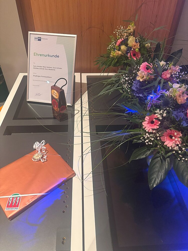 Drei Jahrzehnte voller Wissen und Engagement Zwei bunte Blumenbouquets stehen neben einem eingepackten kleinen Geschenk und einer eingerahmten Ehrenurkunde der IHK für die Euro Akademie und die Euro-Schulen Zwickau.