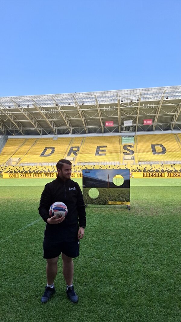 Derselbe Teilnehmer hält den Fußball in der Hand, steht vor der Torwand auf dem Rasen und schaut lächelnd zur Seite, im Hintergrund die riesige „DRESDEN“-Schrift auf der Tribüne.