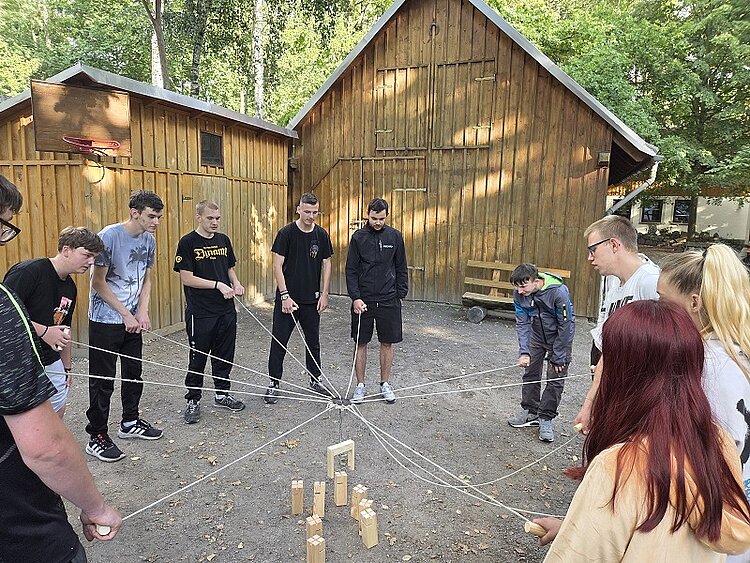 Gruppe Jugendlicher steht im Kreis vor Holzhütten und führt gemeinsam ein Teamspiel mit Seilen und Holzklötzen durch.