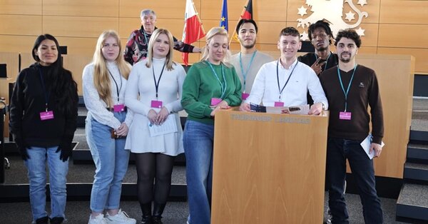 Die Gruppe der Teilnehmenden steht in einem modernen Sitzungssaal des Thüringer Landtages hinter einem Rednerpult. Im Hintergrund sind drei Flaggen zu sehen: eine rot-weiße, eine blaue mit gelben Sternen und eine schwarz-rot-goldene. An der Wand hängt ein großes Wappen mit Löwenmotiv. Die Personen tragen Namensschilder an Bändern um den Hals. Die Sitzreihen im Saal sind leer.