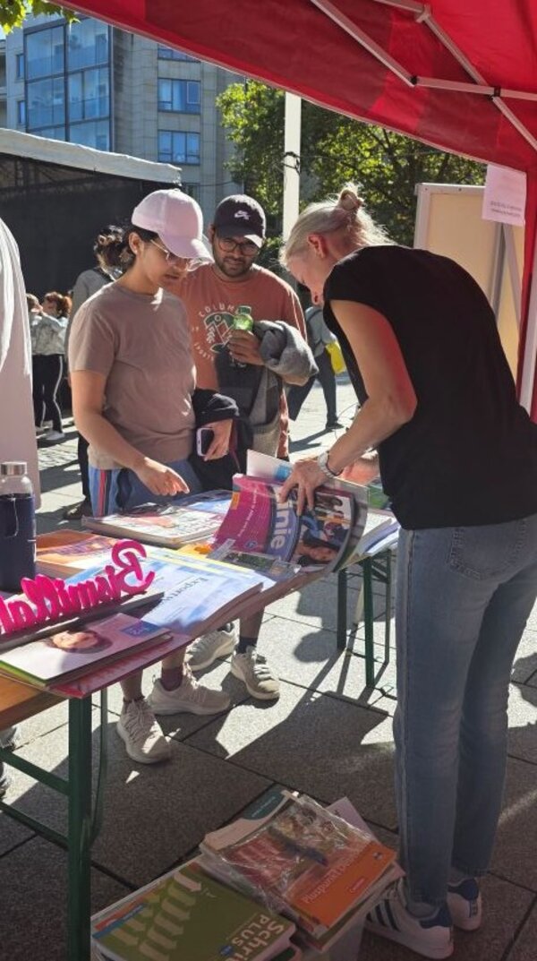 Am Stand der Euro-Schulen unter rotem Sonnenschirm blättern eine Frau und ein Mann in Bücher und Broschüren auf dem mit Materialien gefüllten Tisch; im Hintergrund das Gelände des Straßenfests.