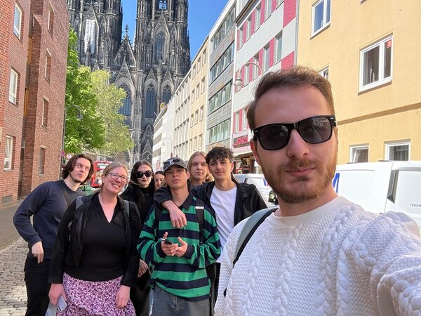 Die Teilnehmergruppe junger Menschen steht auf einer gepflasterten Straße zwischen mehrstöckigen Gebäuden. Im Hintergrund ragt der Kölner Dom mit seinen markanten gotischen Türmen empor. Die Personen tragen legere Kleidung, darunter ein gestreifter Pullover und ein weißer Strickpullover. Ein Teil der Gruppe blickt in die Kamera, während eine Person im Vordergrund ein Selfie aufnimmt. Die Szene ist bei Tageslicht aufgenommen, einige Bäume spenden Schatten.