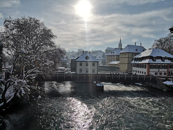 Ein malerischer Winterblick auf einen Fluss in Bamberg. Die Sonne scheint durch einen leicht bewölkten Himmel und spiegelt sich hell auf dem Wasser. Häuser mit schneebedeckten Dächern säumen das Flussufer, und eine Brücke führt über den Fluss. Auch die kahlen Bäume zu beiden Seiten sind mit Schnee bedeckt. Die Szene vermittelt eine friedliche Winteratmosphäre.