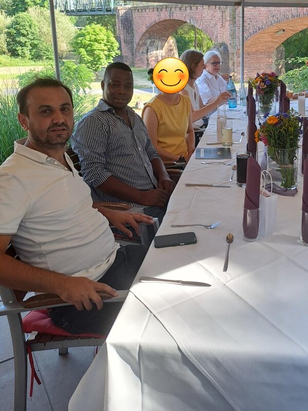 Zwei Männer sitzen an einem langen, weiß gedeckten Tisch auf einer Terrasse mit Blick auf einen Garten und historische Backsteinbögen. Auf dem Tisch stehen Blumen, Gläser und Servietten. Im Hintergrund sind weitere Gäste zu sehen mit Emojis auf ihren Gesichtern. Eine Person hält eine Flasche.