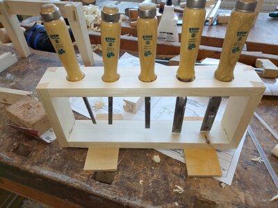 Selbstgebauter Holzständer für Stemmeisen: mehrere Stemmeisen mit Holzgriffen stecken in einer Leiste, darunter hängen die Klingen; das Gestell steht auf einer Werkbank.