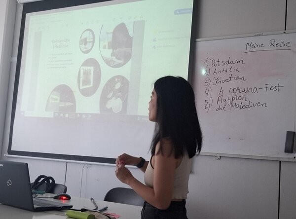 Eine weibliche Teilnehmerin steht vorn am Whiteboard im Unterrichtsraum, sie trägt ein ärmelloses, beiges Top, und hält ihre Präsentation vor ihren Mitteilnehmenden des B2-Sprachkurses an den Euro-Schulen Magdeburg.