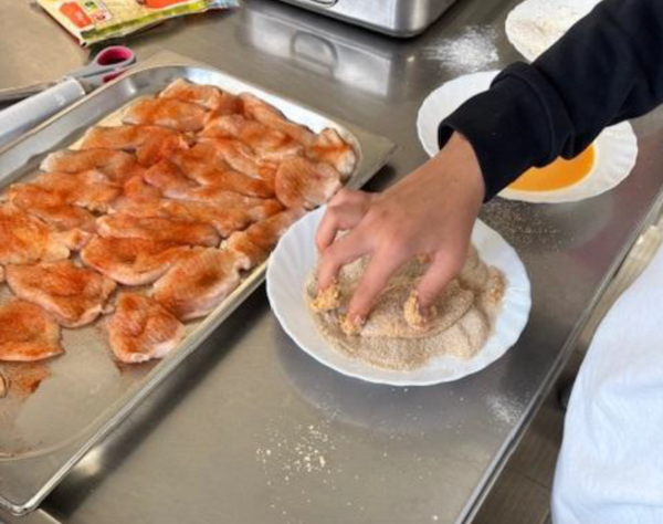 Eine Person paniert ein Stück Fleisch in einer Schale mit Paniermehl. Daneben liegt ein großes Blech mit bereits gewürzten Fleischstücken.