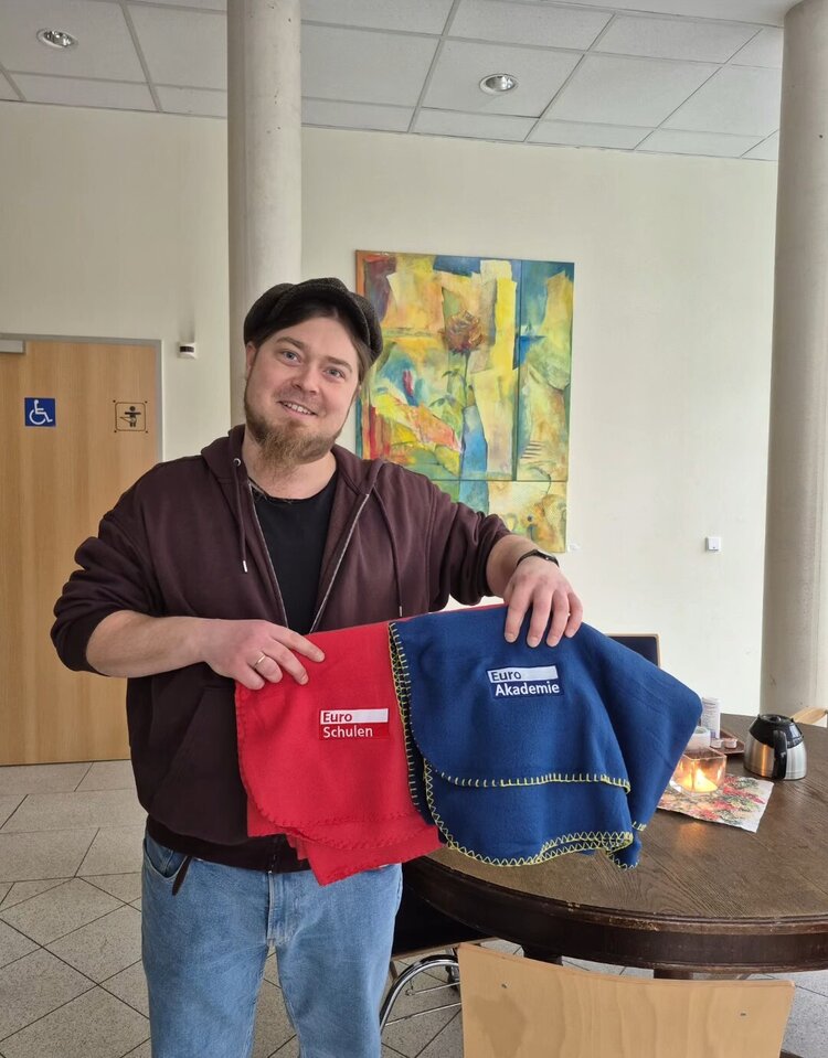 Eine Person steht in einem hellen Innenraum neben einem runden Tisch und hält zwei zusammengefaltete Decken in die Kamera: eine rote mit dem Logo ‚Euro-Schulen‘ und eine blaue mit dem Logo ‚Euro Akademie‘.