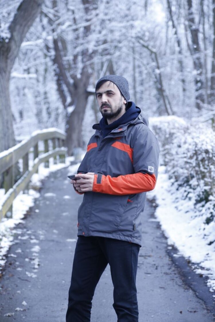 Ein Mann steht auf einem Weg in einer verschneiten Landschaft. Er trägt winterliche Kleidung und hat eine Mütze auf. In den Händen scheint er ein Mobiltelefon zu halten.
