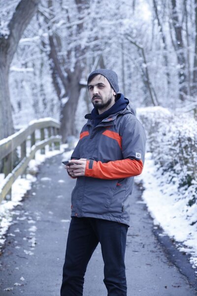 Ein Mann steht auf einem Weg in einer verschneiten Landschaft. Er trägt winterliche Kleidung und hat eine Mütze auf. In den Händen scheint er ein Mobiltelefon zu halten.