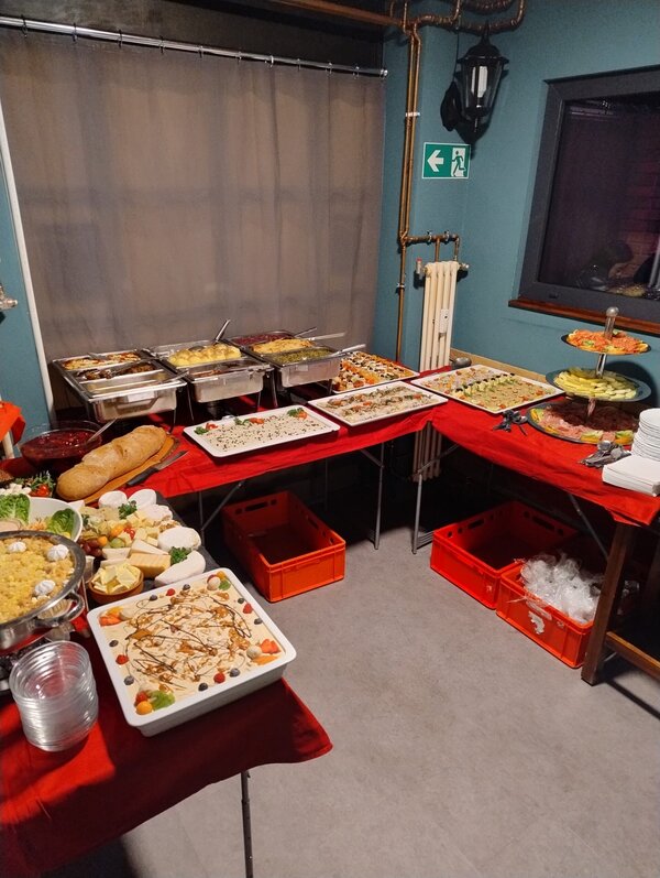 Großes Buffet mit verschiedenen warmen Speisen in Edelstahlbehältern, Platten mit Salaten, Käse, Brot und Desserts, aufgebaut auf rot gedeckten Tischen in einem Gastraum.
