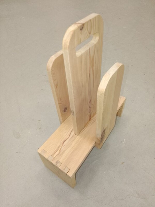 Holz-Zeitungsständer mit drei senkrechten, abgerundeten Seitenteilen und Griffausschnitt; steht auf einer rechteckigen Holz-Basis auf hellem Boden.