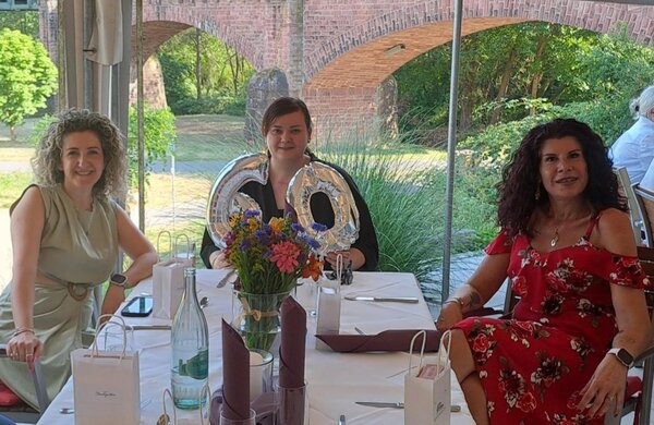 Drei lächelnde Frauen sitzen an einem festlich gedeckten Tisch auf einer Terrasse mit Blick auf einen Garten und historische Backsteinbögen. Auf dem Tisch steht eine silberne '60' als Dekoration sowie ein Blumenstrauß..