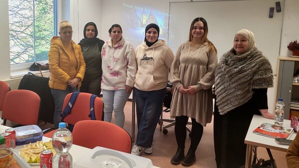 Sechs Frauen aus einem Sprach- oder Integrationskurs stehen nebeneinander im Klassenraum und lächeln in die Kamera, im Hintergrund ist eine Beamerprojektion mit einem Weihnachtsmotiv zu sehen.