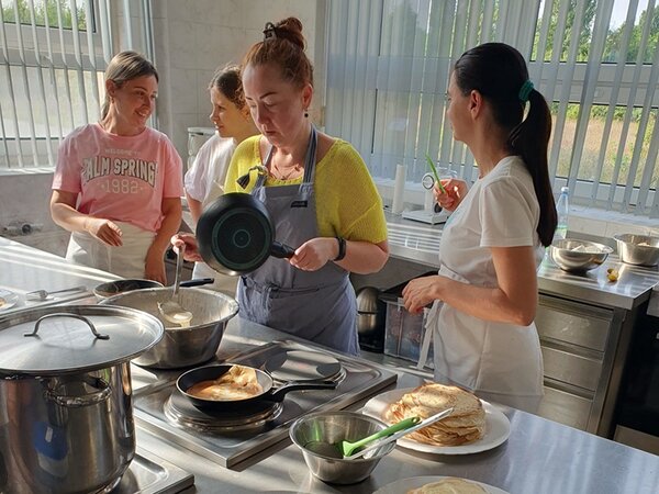 Teilnehmerinnen stehen mit Kochschürzen um den Herd. Es wird sich unterhalten, eine Teilnehmerin rührt den Teig für die vielen Pfannkuchen