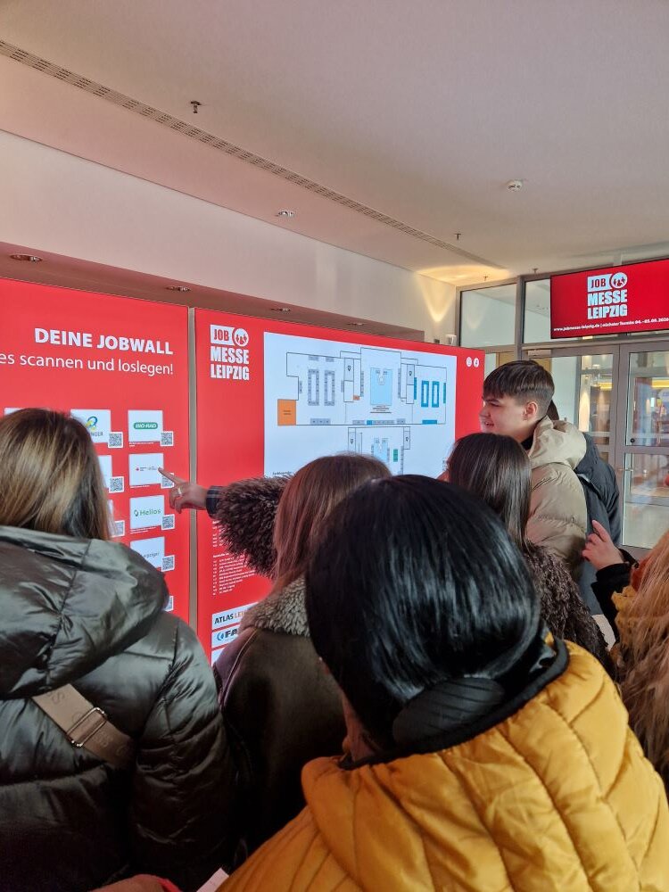 Mehrere Personen stehen vor einer Informationswand. Die Wand ist ebenfrot und zeigt einen Grundriss oder Plan sowie mehrere QR-Codes. Eine Person mit dunkler Jacke zeigt auf einen der QR-Codes, während andere Personen ebenfalls die Inhalte betrachten. Im Hintergrund ist eine Glastür zu sehen, über der "JOB MESSE LEIPZIG" steht.