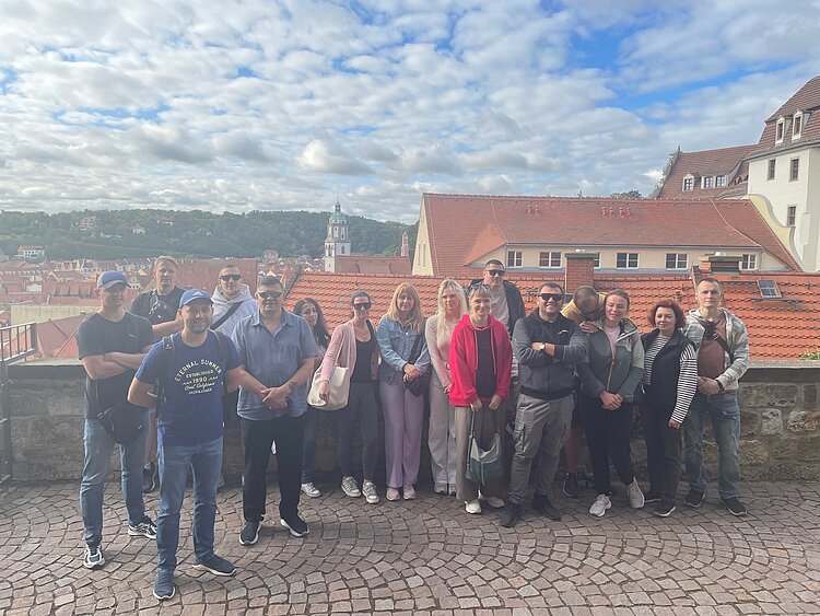Ausflug in Meißen: eine Märchenstadt an der Elbe Eine Gruppe von Menschen steht zusammen auf einem Platz mit Kopfsteinpflaster. Im Hintergrund sind rote Dächer, eine Kirche mit Turm und grüne Hügel unter blauem Himmel mit Wolken zu sehen.