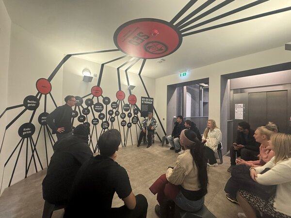 Ein Raum mit heller Beleuchtung, in dem mehrere Teilnehmende auf Hockern und Bänken sitzen und einer stehenden Person zuhören. An der Wand und an der Decke befindet sich eine großflächige, schwarz-rote Grafik mit vielen verbundenen Kreisen, die an ein Netzwerk erinnert. Einige Kreise enthalten weiße Schrift. Die Wände sind hell, der Boden ist gefliest. Im Hintergrund sind eine Tür und ein Aufzug zu sehen.