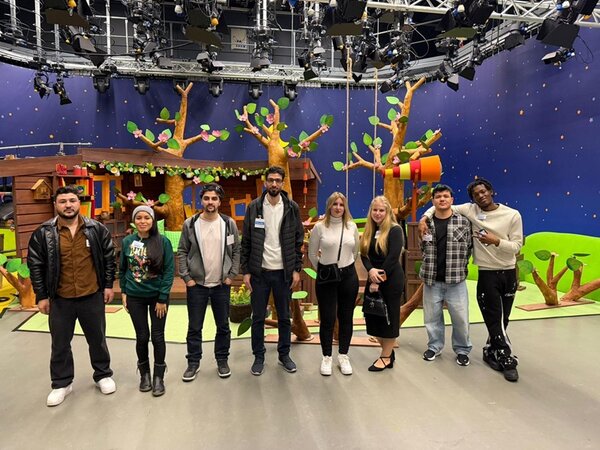 Sieben Teilnehmende stehen nebeneinander im MDR Kinderkanal Fernsehstudio. Im Hintergrund ist eine bunte Kulisse mit künstlichen Bäumen, grünen Blättern und einem Baumhaus zu sehen. Die Wände sind dunkelblau mit gelben Punkten, die an einen Nachthimmel erinnern. Die Personen tragen Freizeitkleidung und blicken zur Kamera. Über ihnen verlaufen zahlreiche Scheinwerfer und technische Geräte an der Studiodecke.