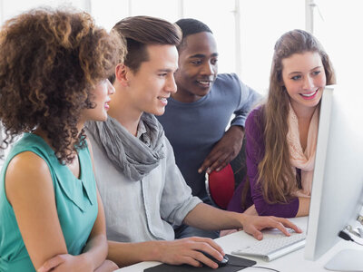 Vier Personen, zwei Frauen und zwei Männer, stehen zusammen vor einem Computer. Sie lächeln und scheinen gemeinsam an etwas auf dem Bildschirm zu arbeiten. Die Szene deutet auf Teamarbeit und Zusammenarbeit in einem Büro- oder Bildungsumfeld hin.